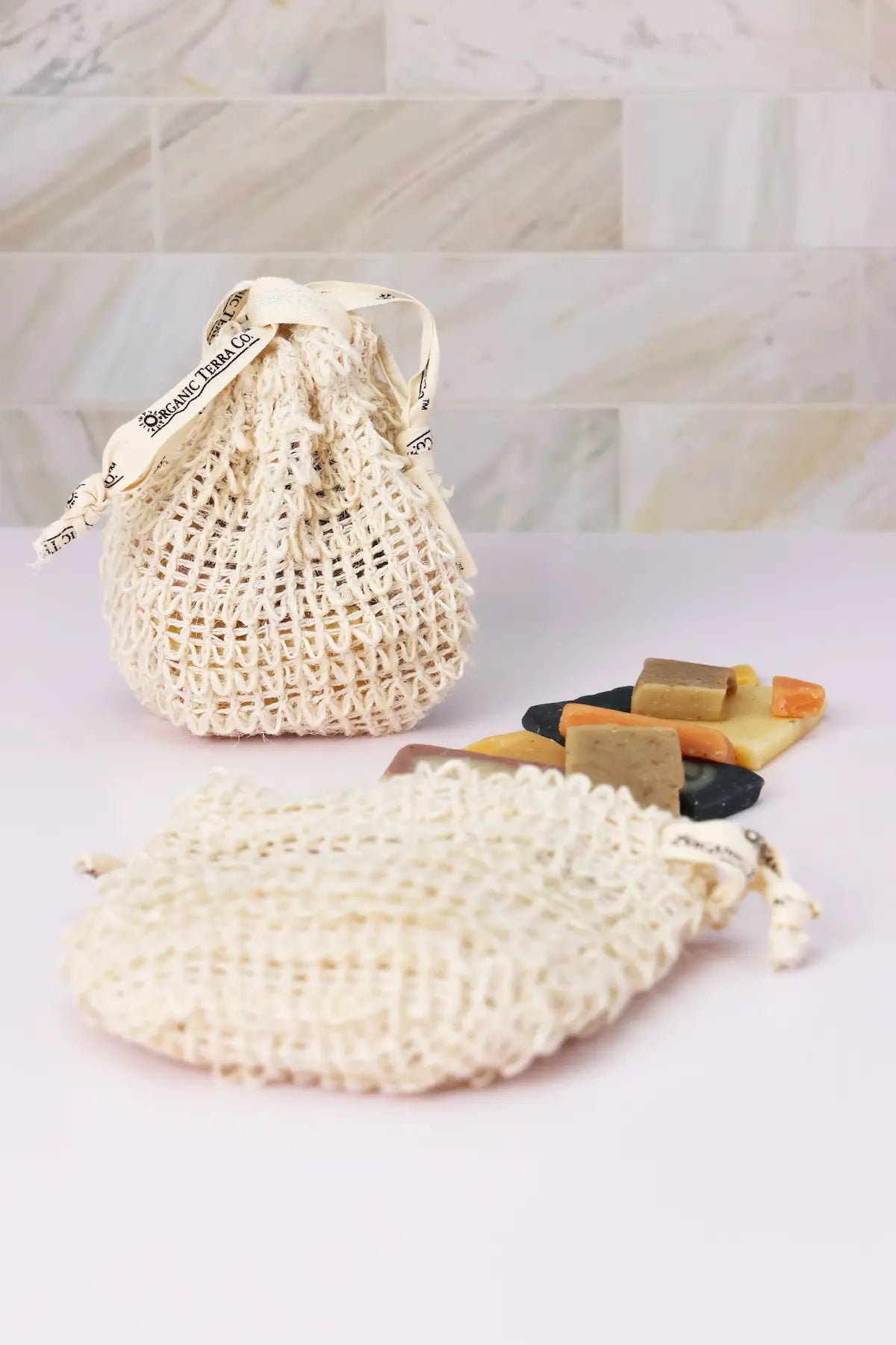 ORGANIC TERRA CO.® sisal drawstring soap pouch and oval sisal soap bag displayed with assorted handcrafted botanical soap bar samples on marble tile surface