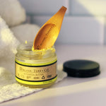 ORGANIC TERRA CO.® engraved wooden spatula resting in open Botanical Butter 2.4 oz glass jar beside folded white towel in bathroom setting