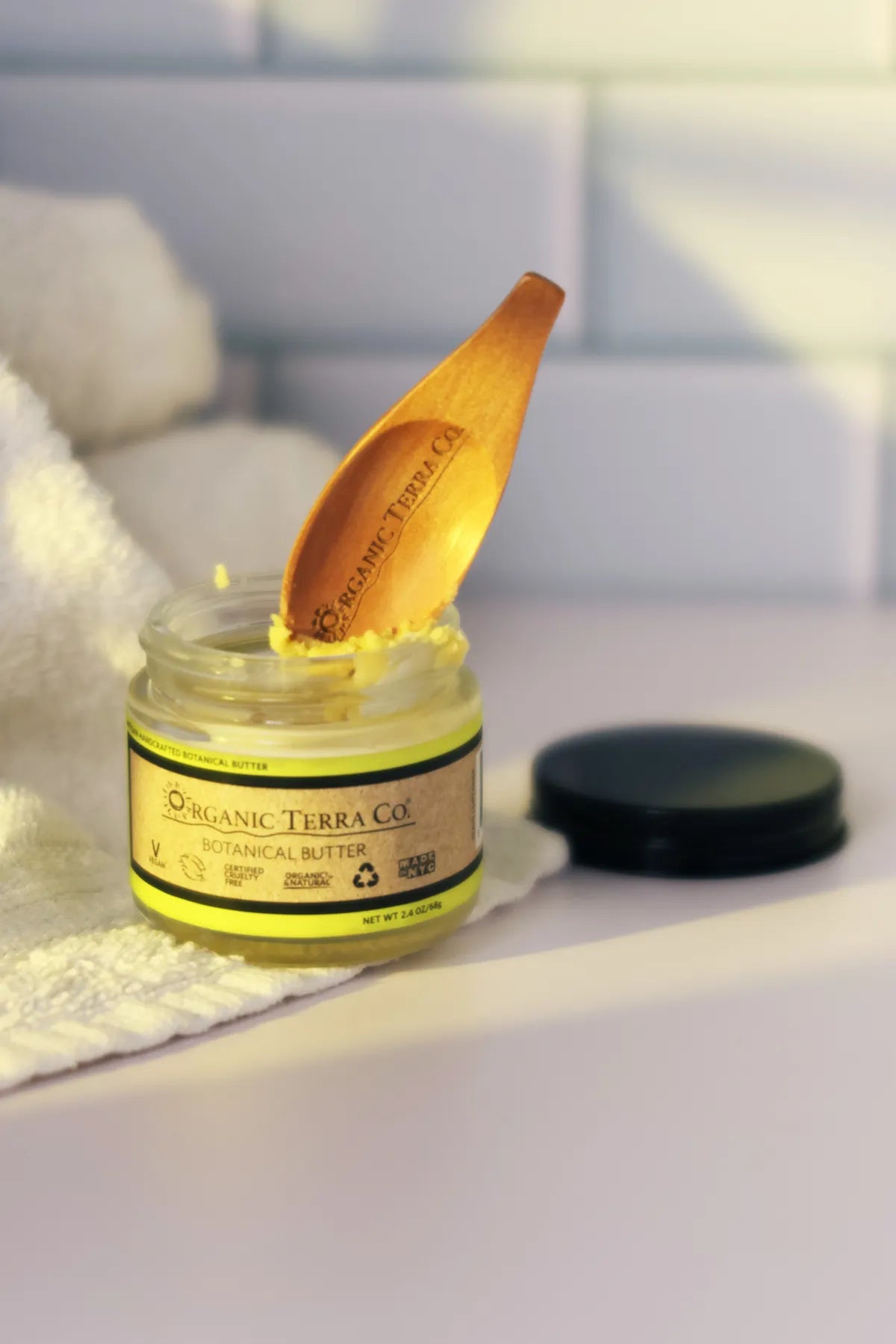 ORGANIC TERRA CO.® engraved wooden spatula resting in open Botanical Butter 2.4 oz glass jar beside folded white towel in bathroom setting