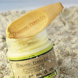 ORGANIC TERRA CO.® wooden spatula resting on open Botanical Butter glass jar nestled in beach sand with ocean in background