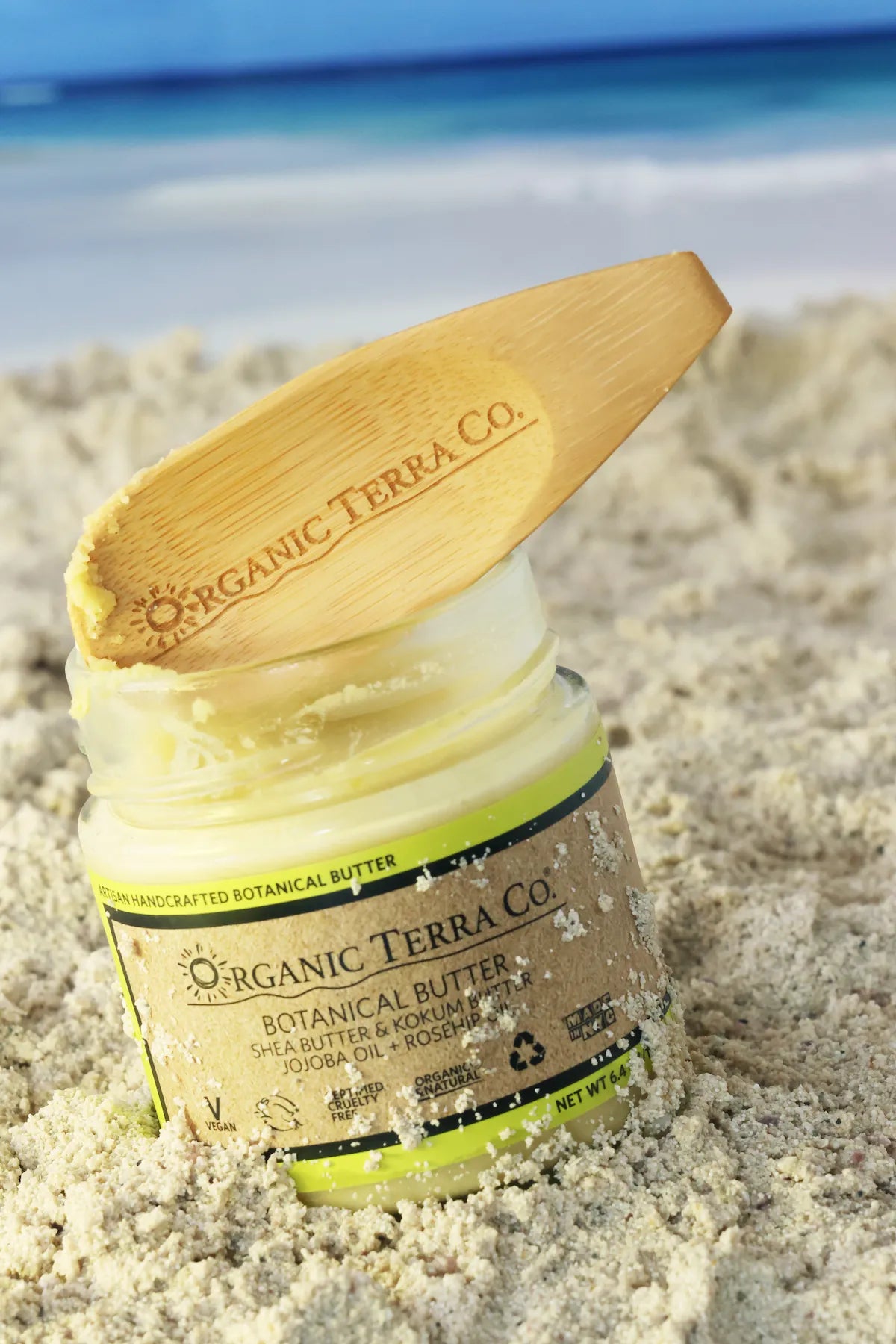 ORGANIC TERRA CO.® wooden spatula resting on open Botanical Butter glass jar nestled in beach sand with ocean in background