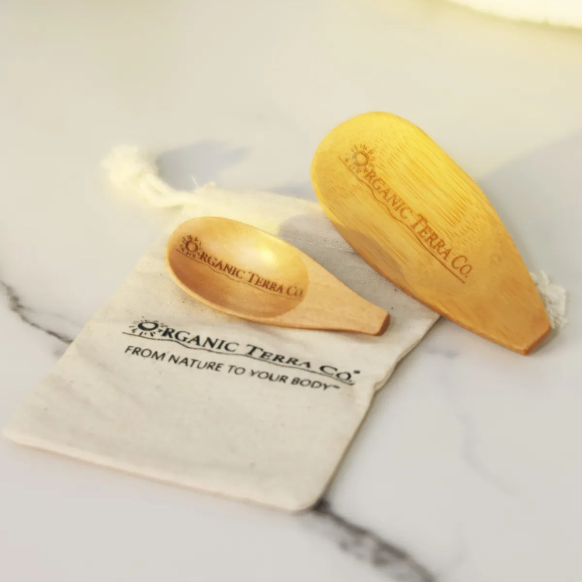 ORGANIC TERRA CO.® engraved wooden spoon and wooden spatula on branded cotton muslin pouch displaying From Nature To Your Body trademark on marble surface