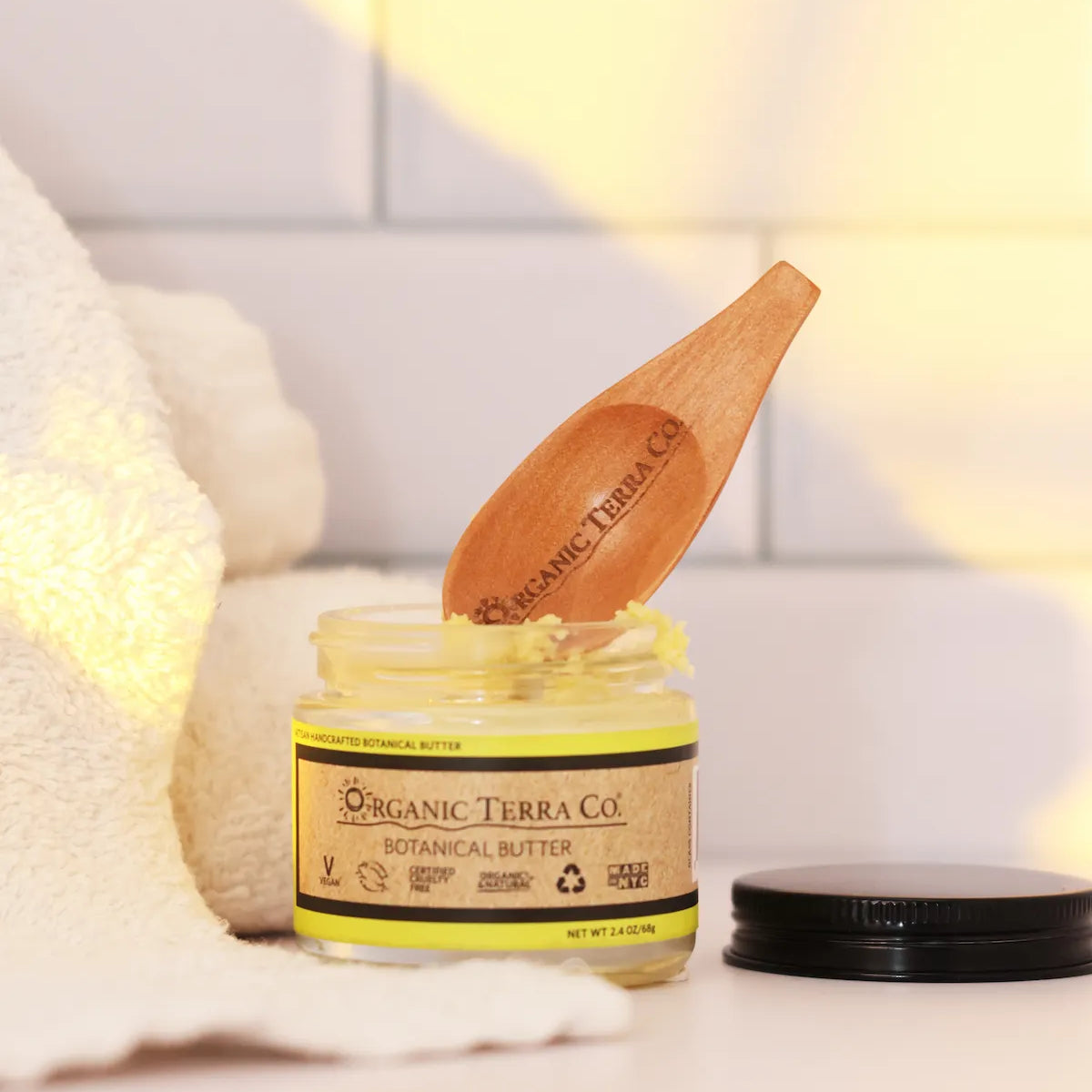 ORGANIC TERRA CO.® Botanical Butter 2.4 oz open glass jar with engraved wooden spatula resting on jar beside folded towel in warm bathroom setting