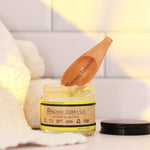 ORGANIC TERRA CO.® Botanical Butter 2.4 oz open glass jar with engraved wooden spatula resting on jar beside folded towel in warm bathroom setting