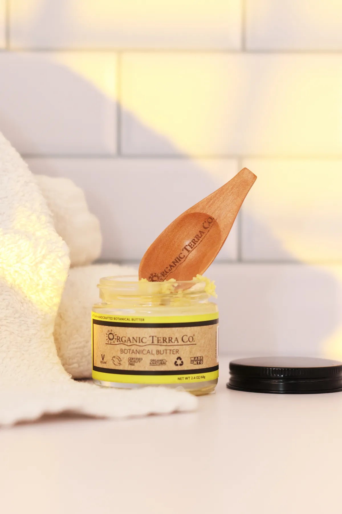 ORGANIC TERRA CO.® Botanical Butter 2.4 oz open glass jar with engraved wooden spatula resting on jar beside folded towel in warm bathroom setting
