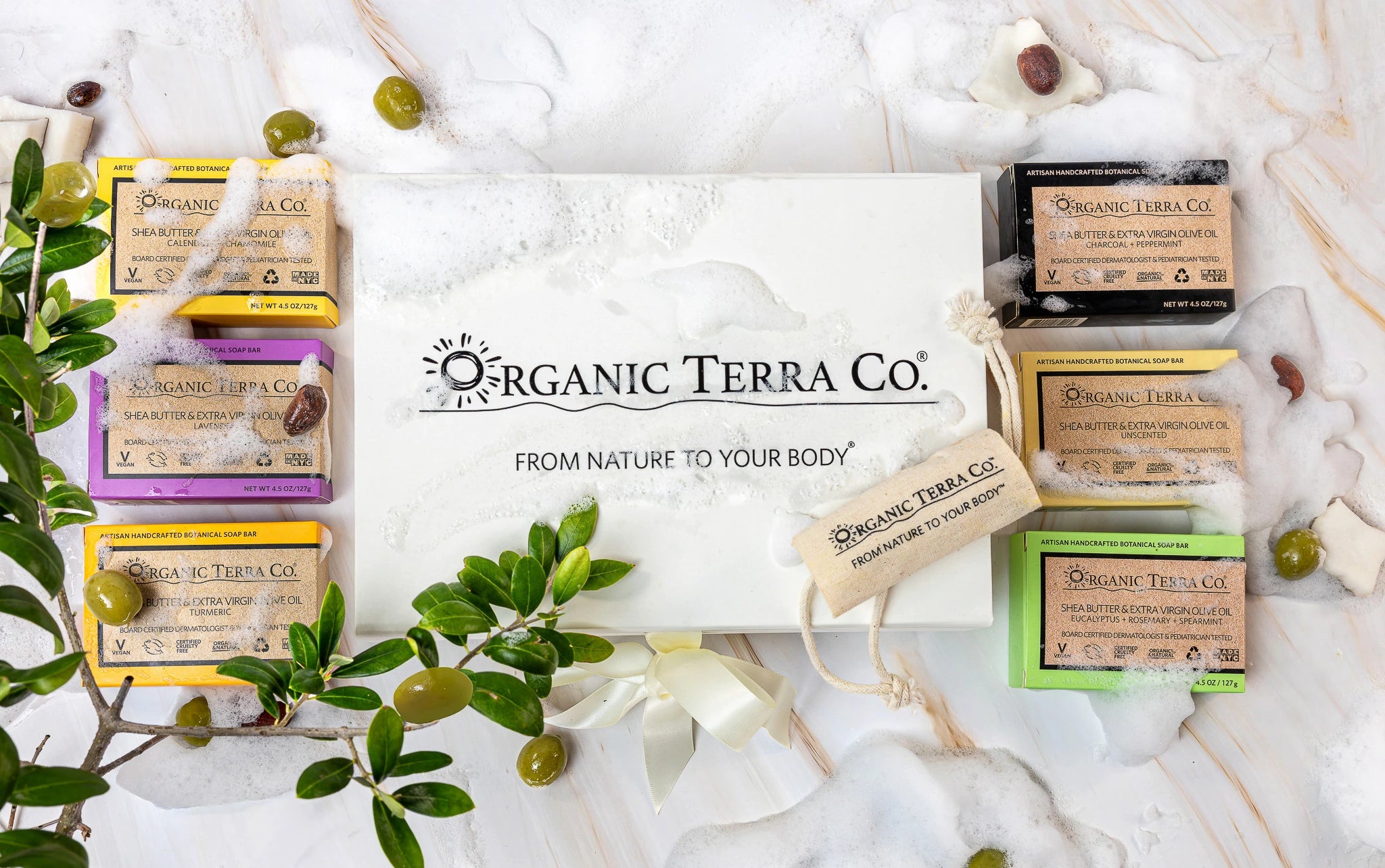Organic Terra Co. Botanical Cleansing Collection gift set displayed on white marble with six artisan soap bars, olive branches, and soap suds.