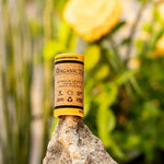 ORGANIC TERRA CO.® Botanical Lip Balm tube balanced on natural rock surrounded by lush green plants and yellow flowers in botanical garden setting
