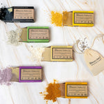 Six Organic Terra Co. botanical soap bars arranged on marble, each beside its key natural ingredient — charcoal, turmeric, lavender, eucalyptus, and calendula powders.
