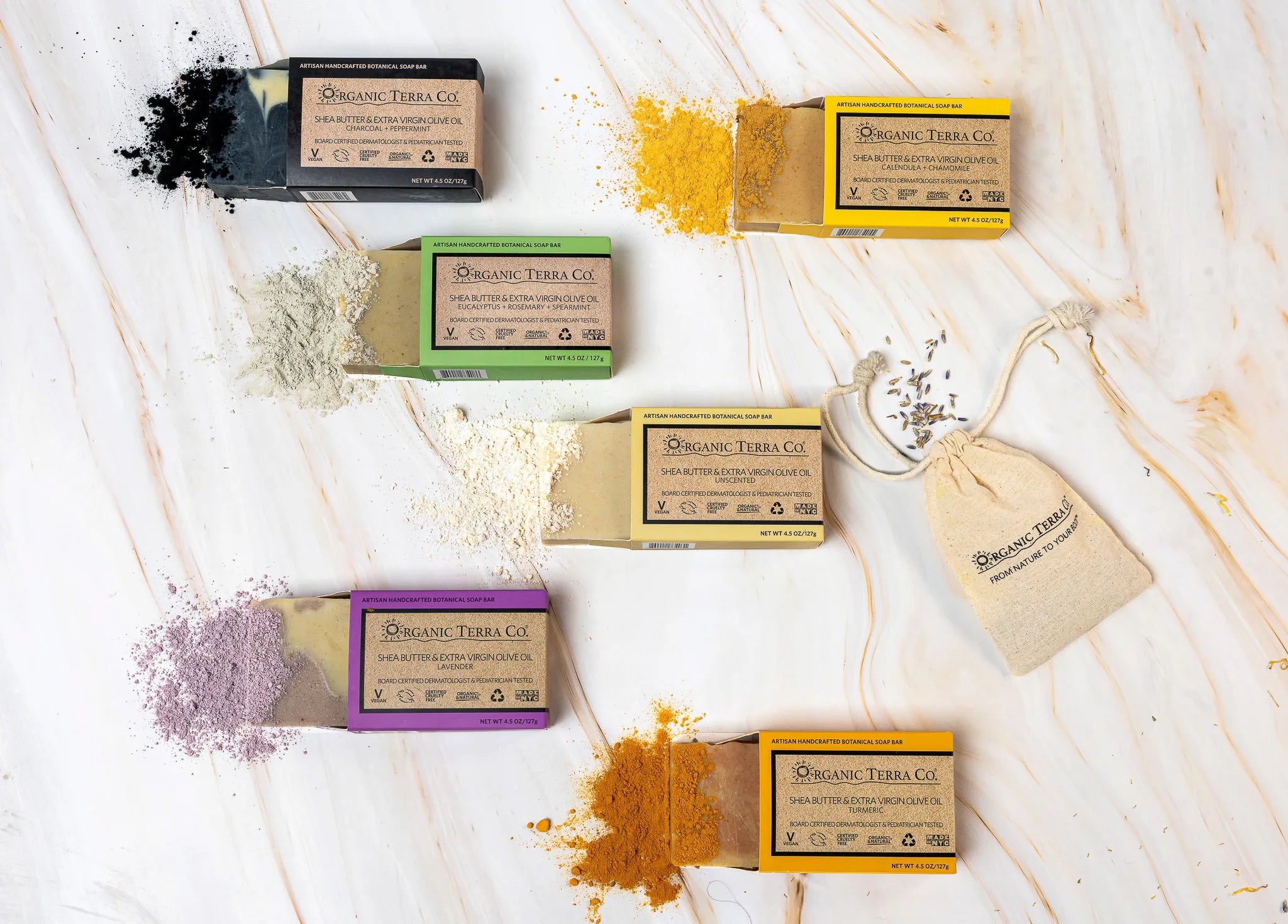 Six Organic Terra Co. botanical soap bars arranged on marble, each beside its key natural ingredient — charcoal, turmeric, lavender, eucalyptus, and calendula powders.