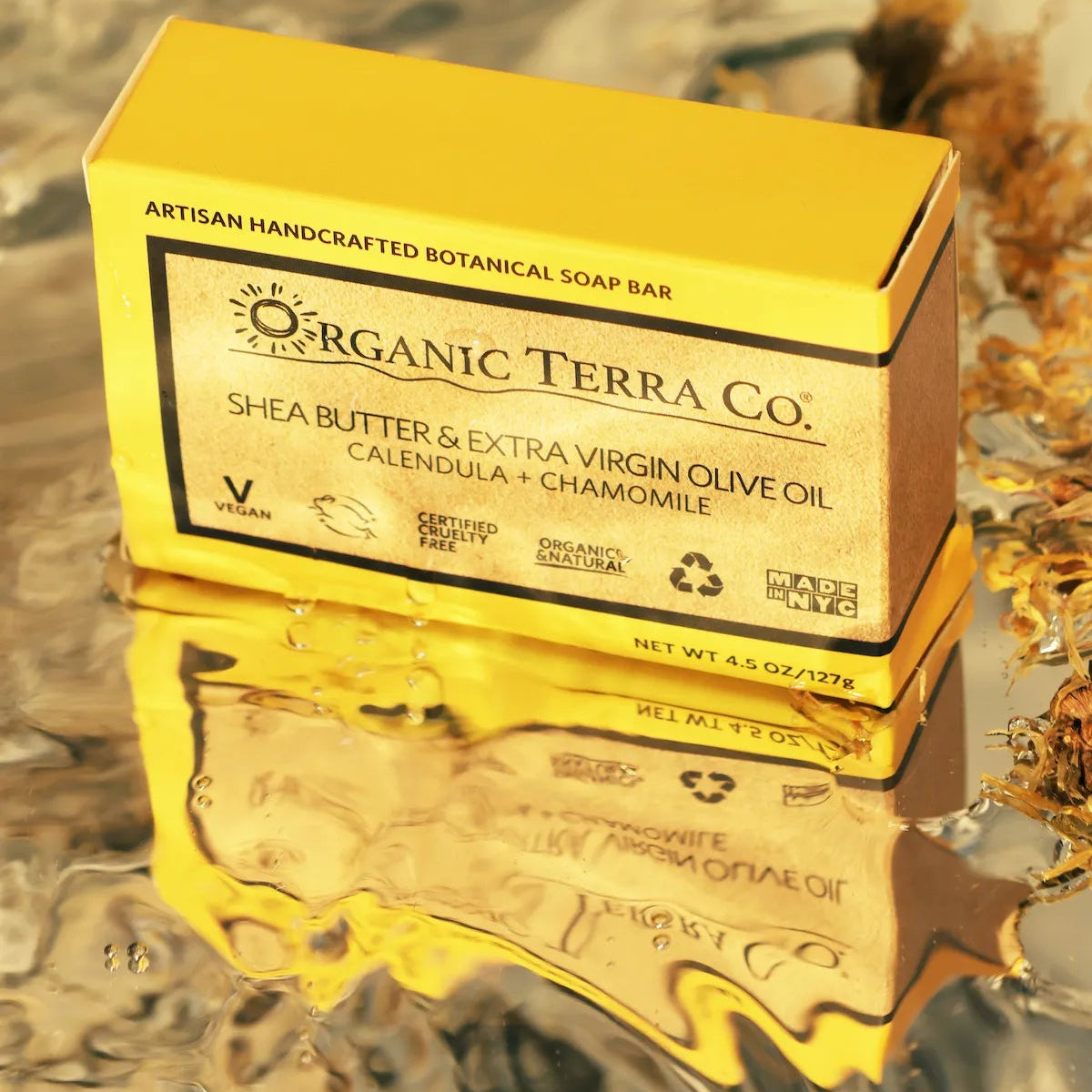 .ORGANIC TERRA CO.®Shea Butter Extra Virgin Olive Oil Calendula and Chamomile soap bar box reflected on wet surface beside dried calendula petals