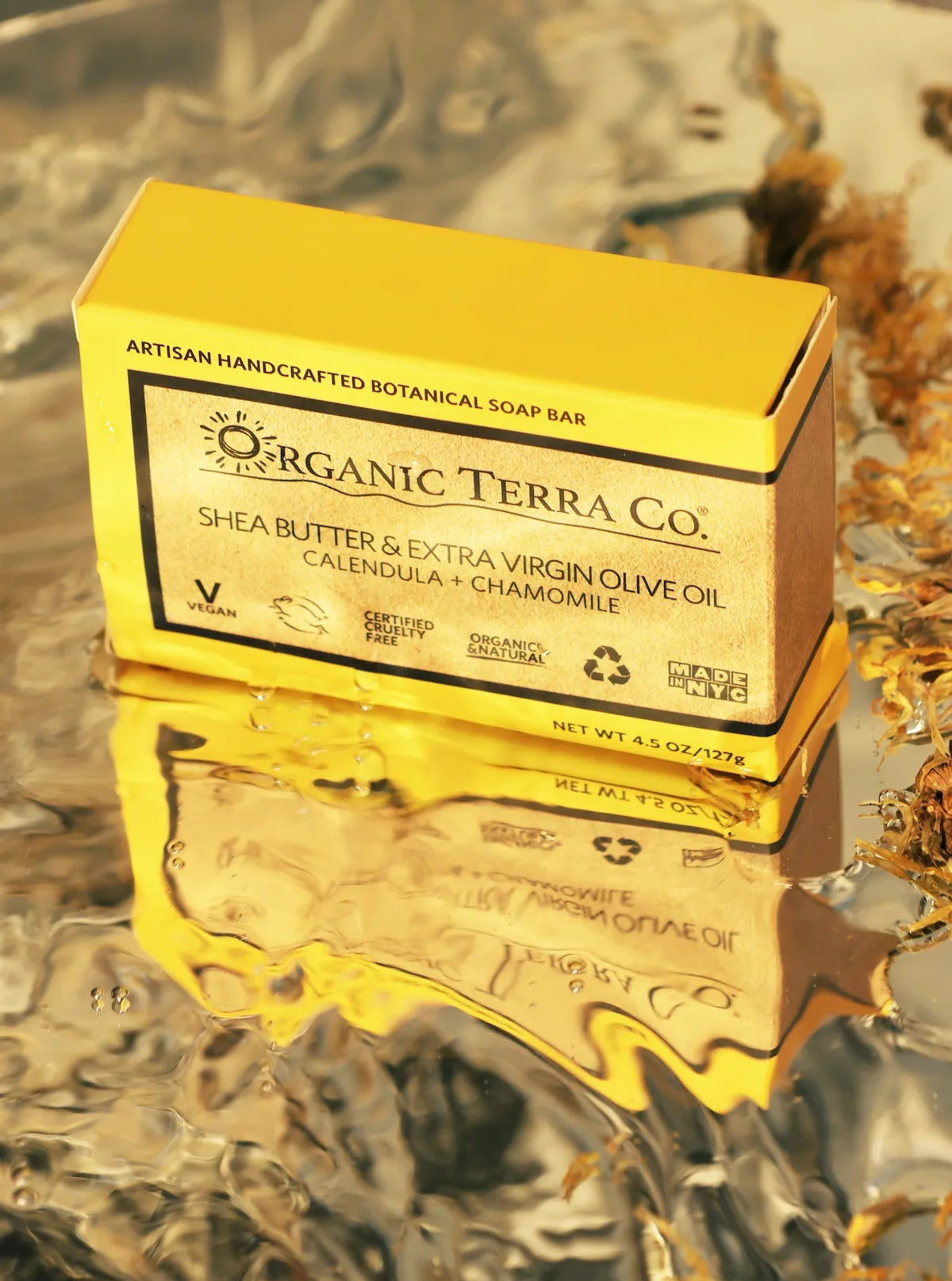 .ORGANIC TERRA CO.®Shea Butter Extra Virgin Olive Oil Calendula and Chamomile soap bar box reflected on wet surface beside dried calendula petals
