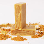 Two ORGANIC TERRA CO.® Shea Butter Extra Virgin Olive Oil Calendula and Chamomile soap bars stacked surrounded by turmeric powder and dried calendula on white background