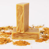 Two ORGANIC TERRA CO.® Shea Butter Extra Virgin Olive Oil Calendula and Chamomile soap bars stacked surrounded by turmeric powder and dried calendula on white background