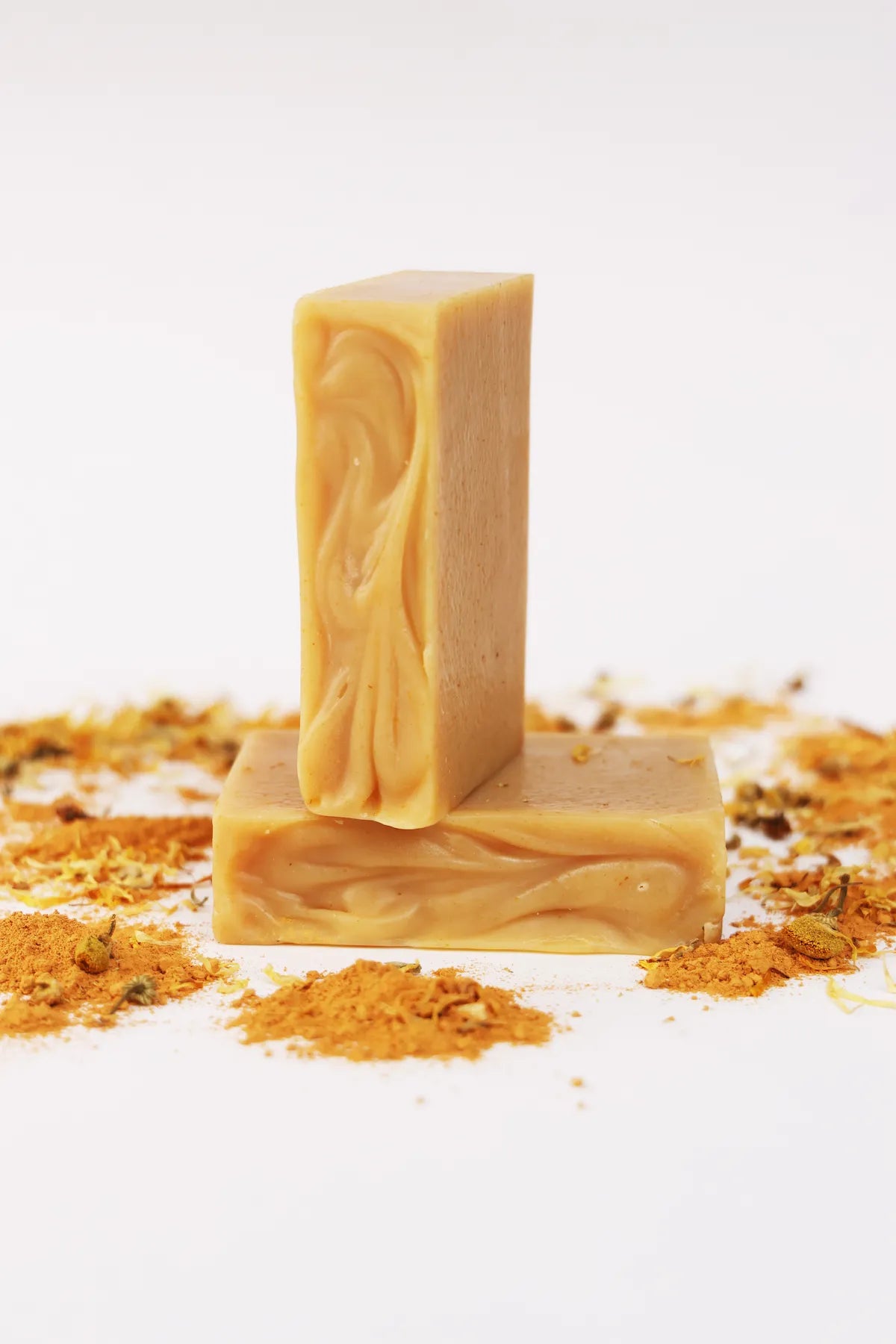 Two ORGANIC TERRA CO.® Shea Butter Extra Virgin Olive Oil Calendula and Chamomile soap bars stacked surrounded by turmeric powder and dried calendula on white background