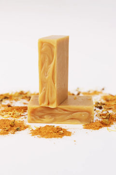 Two ORGANIC TERRA CO.® Shea Butter Extra Virgin Olive Oil Calendula and Chamomile soap bars stacked surrounded by turmeric powder and dried calendula on white background