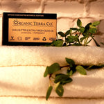 ORGANIC TERRA CO.® Butter Extra Virgin Olive Oil Charcoal and Peppermint soap bar box resting on stacked white towels with fresh peppermint sprigs against white brick wall in warm light