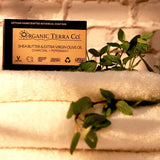 ORGANIC TERRA CO.® Butter Extra Virgin Olive Oil Charcoal and Peppermint soap bar box resting on stacked white towels with fresh peppermint sprigs against white brick wall in warm light