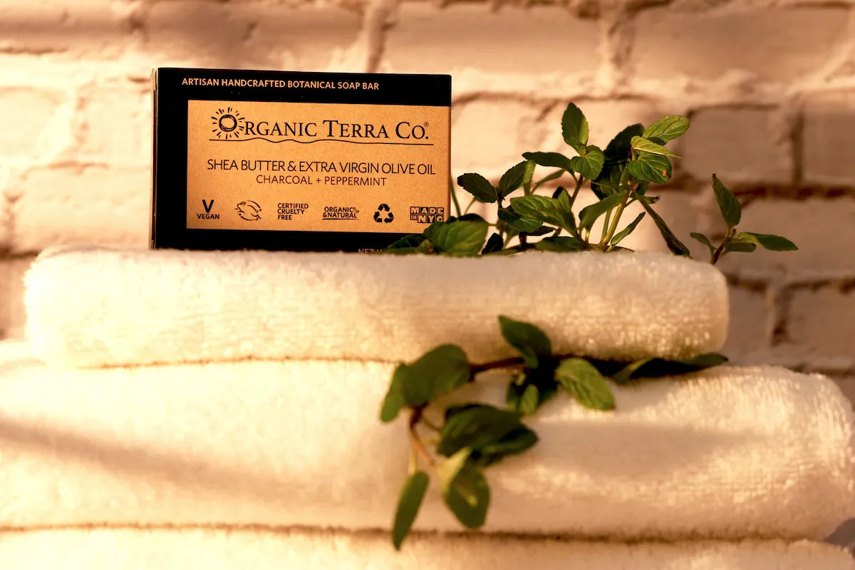 ORGANIC TERRA CO.® Butter Extra Virgin Olive Oil Charcoal and Peppermint soap bar box resting on stacked white towels with fresh peppermint sprigs against white brick wall in warm light