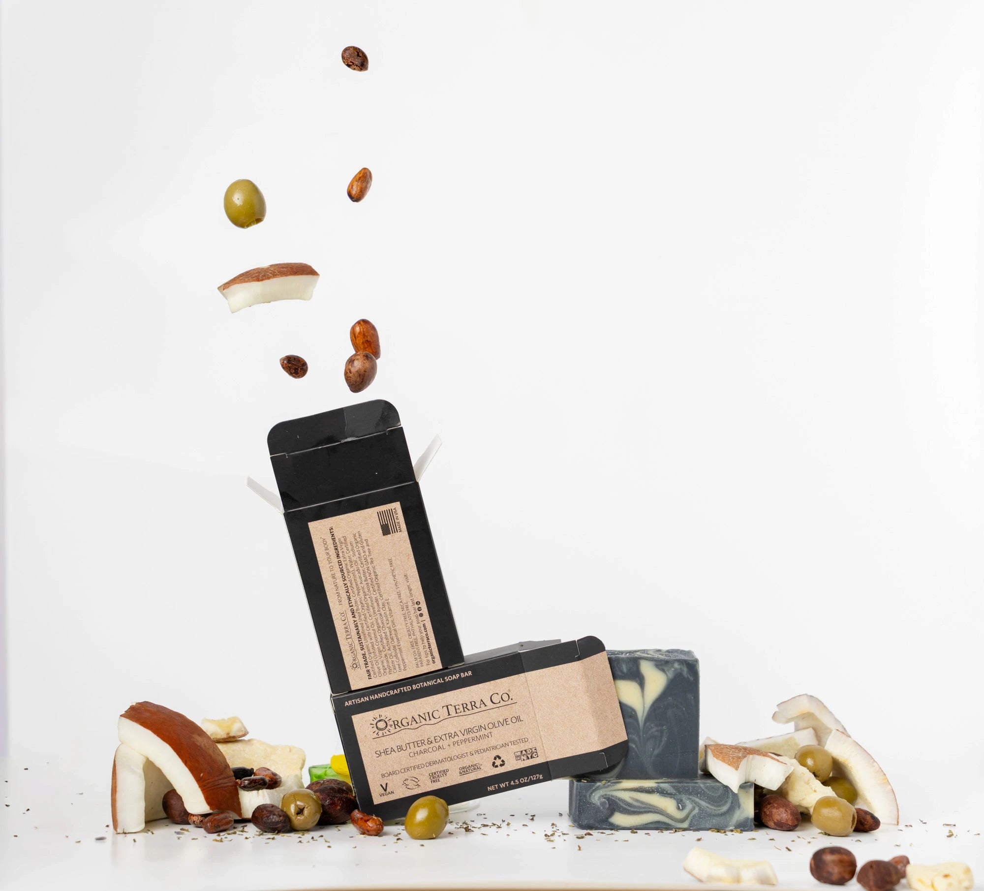 ORGANIC TERRA CO.®Shea Butter Extra
Virgin Olive Oil Charcoal and Peppermint soap bar front and back packaging with unwrapped bars and scattered shea nuts olives and cacao beans