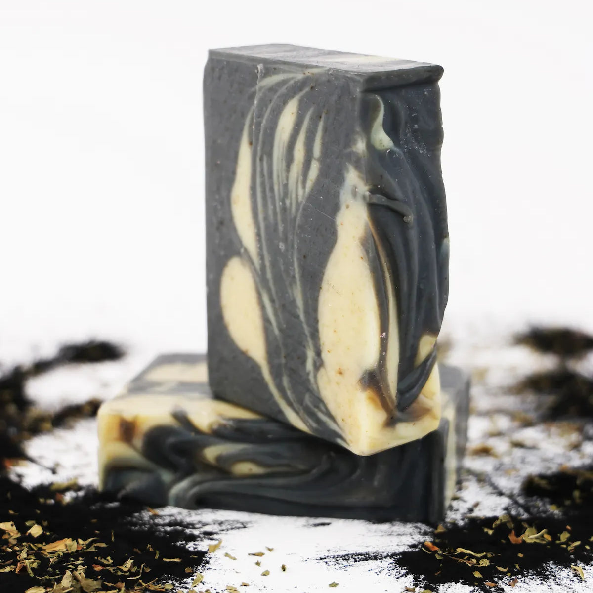 Two ORGANIC TERRA CO.®handcrafted Shea Butter Extra Virgin Olive Oil Charcoal and Peppermint soap bars stacked surrounded by activated charcoal powder and dried herbs on white background