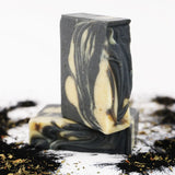 Two ORGANIC TERRA CO.®handcrafted Shea Butter Extra Virgin Olive Oil Charcoal and Peppermint soap bars stacked surrounded by activated charcoal powder and dried herbs on white background
