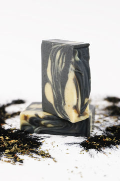 Two ORGANIC TERRA CO.®handcrafted Shea Butter Extra Virgin Olive Oil Charcoal and Peppermint soap bars stacked surrounded by activated charcoal powder and dried herbs on white background