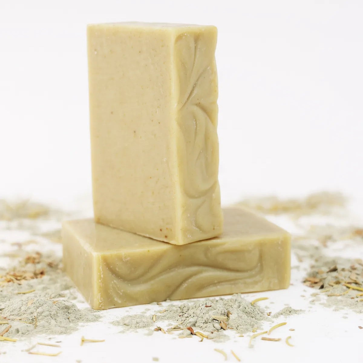 Two ORGANIC TERRA CO.®handcrafted Shea Butter Extra Virgin Olive Oil Eucalyptus Rosemary and Spearmint soap bars stacked surrounded by french green clay powder and dried herbs on white background