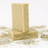Two ORGANIC TERRA CO.®handcrafted Shea Butter Extra Virgin Olive Oil Eucalyptus Rosemary and Spearmint soap bars stacked surrounded by french green clay powder and dried herbs on white background