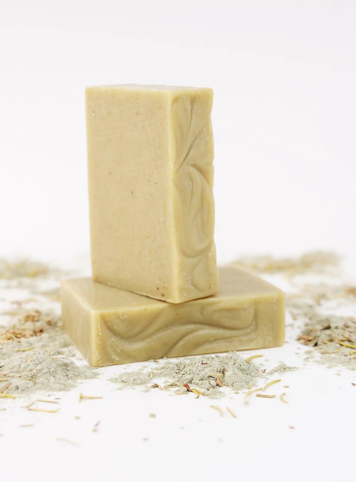 Two ORGANIC TERRA CO.®handcrafted Shea Butter Extra Virgin Olive Oil Eucalyptus Rosemary and Spearmint soap bars stacked surrounded by french green clay powder and dried herbs on white background