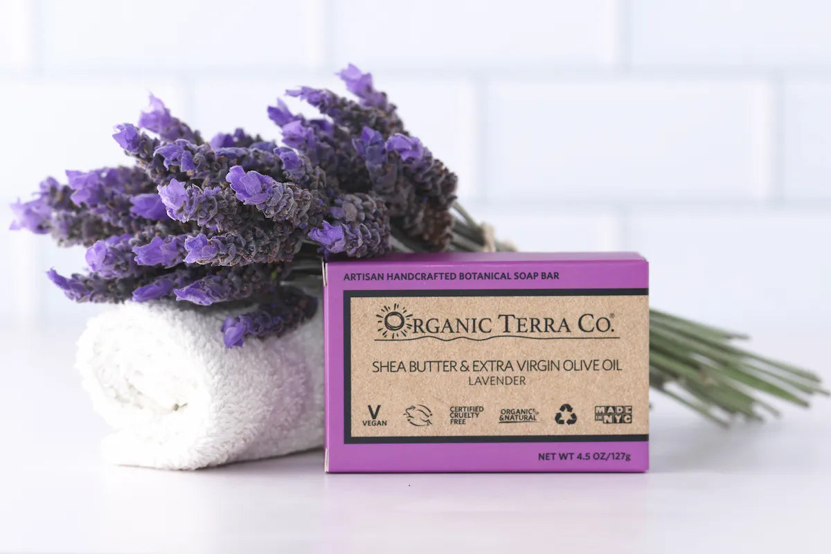 ORGANIC TERRA CO.®Shea Butter Extra Virgin Olive Oil Lavender soap bar box beside rolled white towel and large fresh lavender bouquet against white tile background