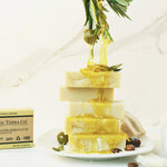 Stack of ORGANIC TERRA CO.®Shea Butter and Extra Virgin Olive Oil unscented handcrafted soap bars with olive oil dripping from olive branch on marble surface beside product box