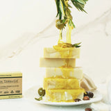 Stack of ORGANIC TERRA CO.®Shea Butter and Extra Virgin Olive Oil unscented handcrafted soap bars with olive oil dripping from olive branch on marble surface beside product box