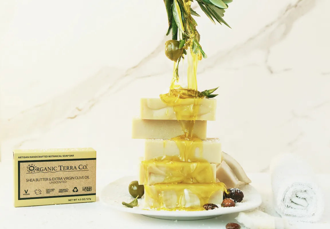 Stack of ORGANIC TERRA CO.®Shea Butter and Extra Virgin Olive Oil unscented handcrafted soap bars with olive oil dripping from olive branch on marble surface beside product box