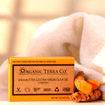 ORGANIC TERRA CO.® Butter Extra Virgin Olive Oil Turmeric soap bar box beside white towel and fresh sliced turmeric root in warm bathroom light