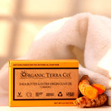ORGANIC TERRA CO.® Butter Extra Virgin Olive Oil Turmeric soap bar box beside white towel and fresh sliced turmeric root in warm bathroom light