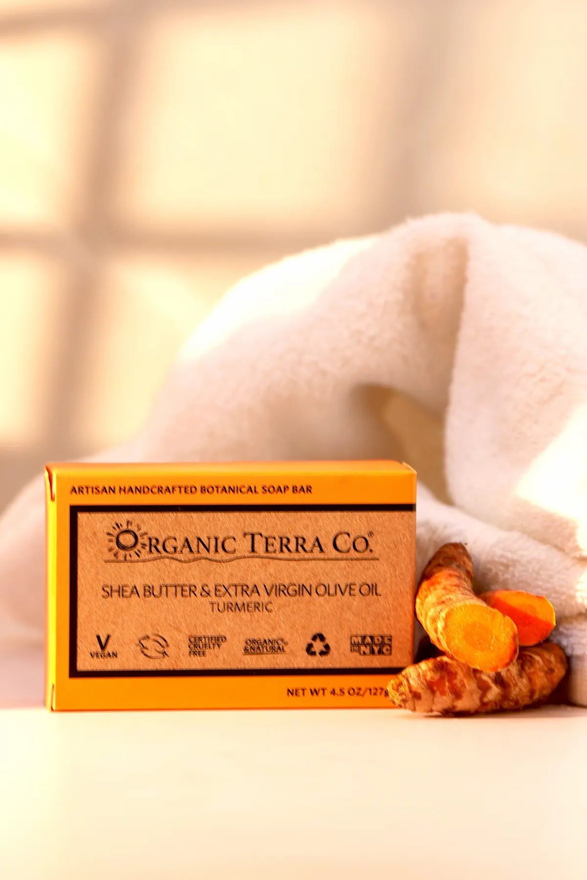 ORGANIC TERRA CO.® Butter Extra Virgin Olive Oil Turmeric soap bar box beside white towel and fresh sliced turmeric root in warm bathroom light